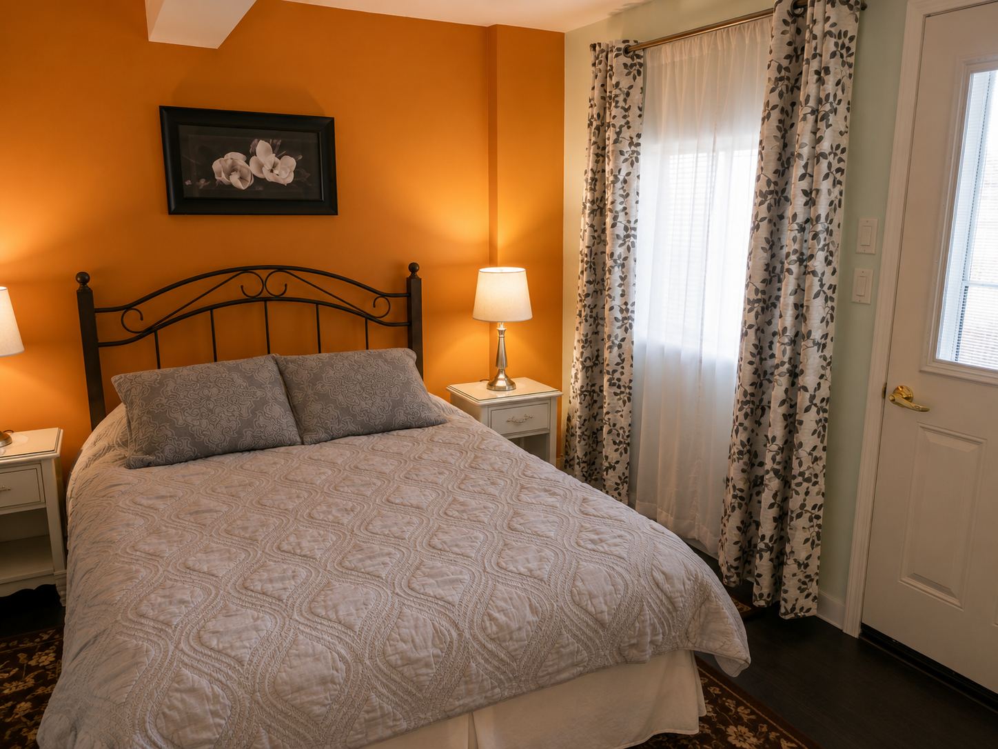 The Blue Heron suite: a queen bed against a warm orange wall, a lamp on the night table and a curtained door to the outside.