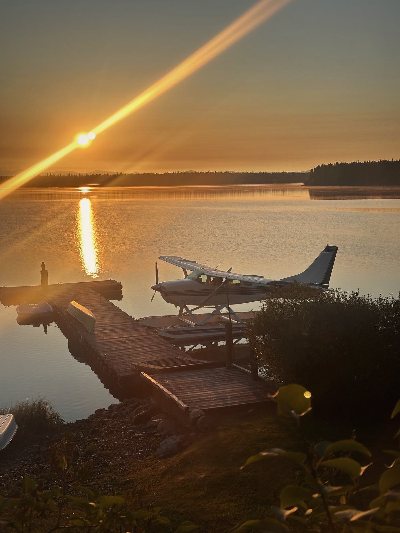 A float-equipped Cessna tied at the Eagle's Nest dock at sunrise, the sun low over Anahim Lake and a canoe and pedal boats on the dock behind it.