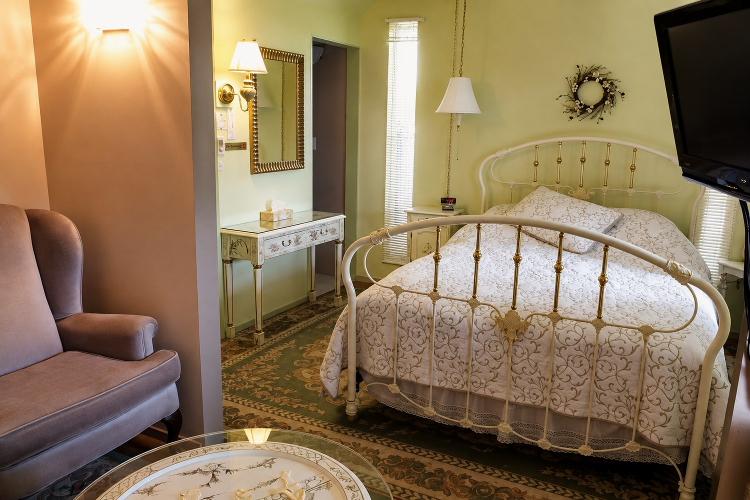 The Green suite: a queen bed in a white and brass frame with a cream cover, pale green walls, and an armchair in the foreground.