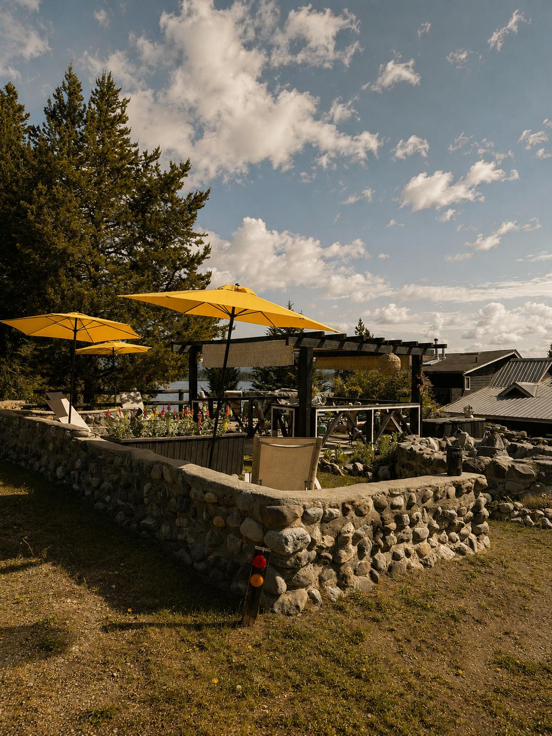 The Guest Garden — a river-stone wall, flower beds, a pergola and yellow umbrellas above tables looking out to the lake.