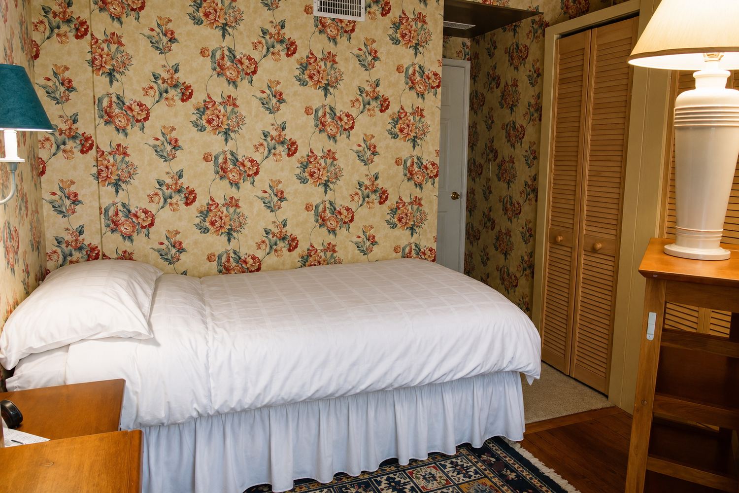 The Pelican room: a bed made up in white against floral papered walls, with a louvred wardrobe and a lamp on the side table.