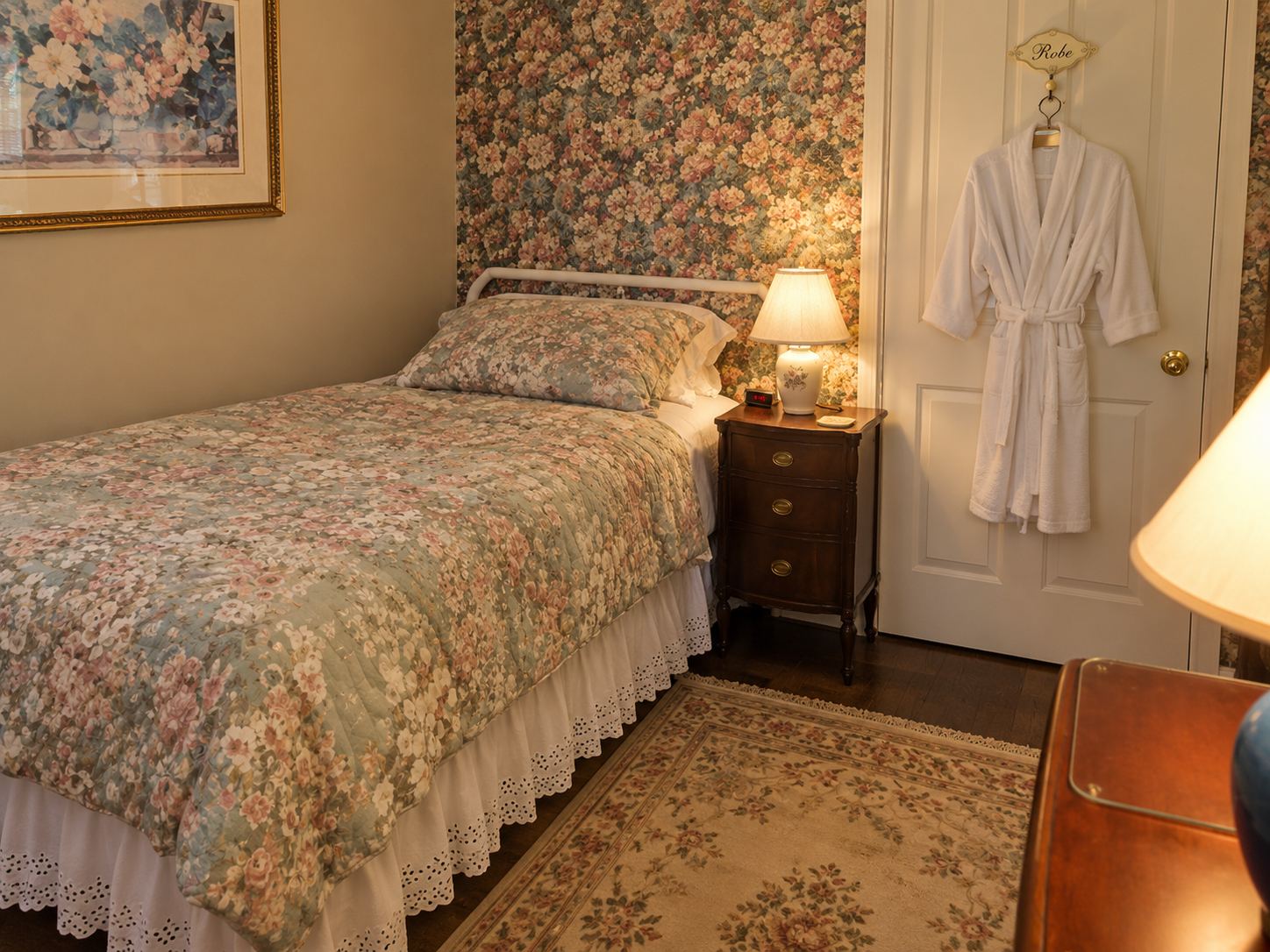 The Pine room: a twin bed with a floral quilt against patterned wallpaper, a lamp lit on the bedside table and a robe hanging on the door.